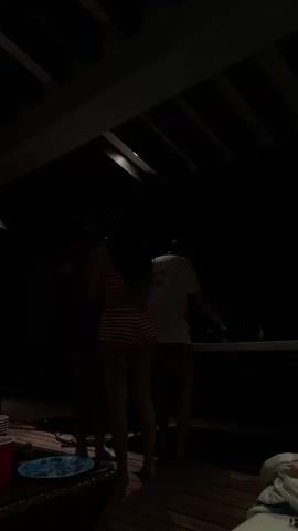 The video shows a group of people enjoying themselves at what appears to be a nighttime gathering. 

Initially, three individuals are dancing on a wooden deck, their movements indistinct due to the low lighting. There's a table nearby with cups, plates, and other party items.

The video then shifts focus, revealing a man in an open robe walking into view. In the foreground, several people are reclining, with their legs and feet visible. A few plastic cups are scattered around the lounging area. 

The scene suggests a casual, relaxed atmosphere, possibly a party or get-together in a private residence.