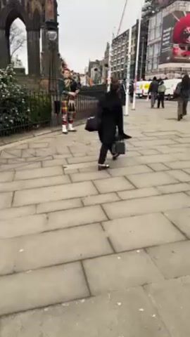The video shows a man playing the bagpipes on a city sidewalk in front of a wrought iron fence. He is wearing a traditional Scottish kilt with a green vest and white hose.  A black bag is open on the sidewalk nearby, presumably to collect money. People walk past the bagpiper. The background includes a large building, what appears to be a historical church ruin, and a street with cars and pedestrian traffic. The weather appears overcast.