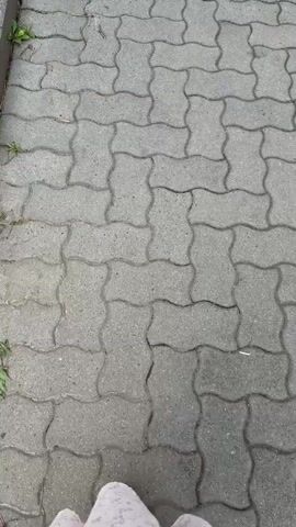 A first-person perspective video shows someone walking on a paved surface. The ground is made of interlocking gray concrete pavers arranged in a wavy pattern. A single person's feet and legs are visible in the frame. They are wearing gray sneakers with white laces and a white skirt with a subtle pattern. The person is moving forward, indicating they are walking. The environment appears to be outdoors, possibly a sidewalk or a pedestrian path. The lighting suggests it is daytime.
