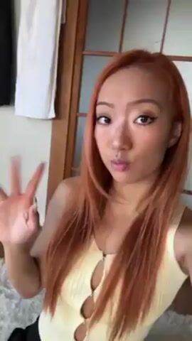 A young woman with reddish-brown, long straight hair is holding a phone camera at arm's length, and is pointing it at herself. She is sitting on a fluffy white carpet in a room with a white wall, a light-colored wood floor, and a light-colored wood sliding door. She is wearing a yellow tank top with a zipper and cut-outs on the chest, and black pants. She waves at the camera with her right hand, and looks at the camera. A pair of black pants is hanging from the corner of a wall.