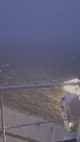 A first-person perspective video shows a ship sailing through dark, choppy water during a snowstorm. The camera is positioned on the deck, looking out over the railing. Snowflakes are visible falling and being blown by the wind, creating a hazy effect on the water. The water itself appears rough, with whitecaps and foam indicating choppy conditions. A railing runs across the bottom of the frame, and a white object, possibly a light fixture or piece of equipment, is mounted on it. The lighting is dim, with the only significant light source appearing to be this mounted fixture. The overall impression is one of a challenging maritime environment.