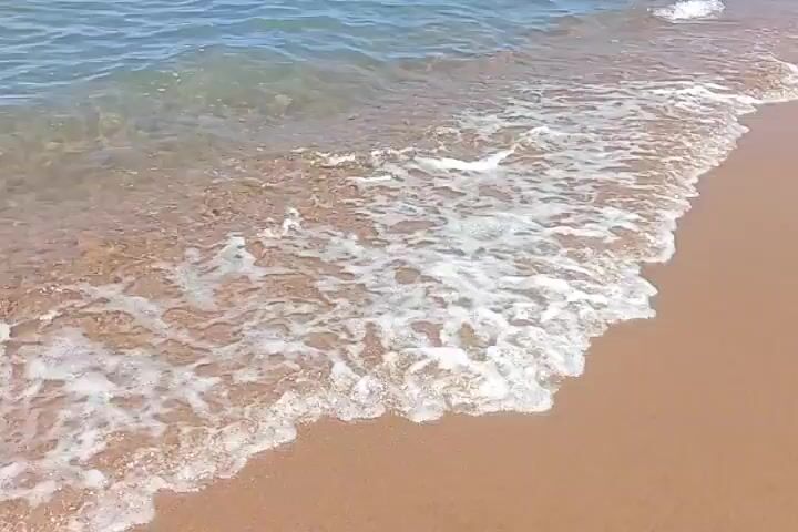 The video shows a close-up view of gentle waves lapping onto a sandy beach. The water is clear and shallow, revealing the sand beneath. The sand is a light brown color. The waves create foamy white edges as they reach the shore, then recede, leaving a glistening wet surface. The horizon is visible in the distance, with a hint of blue sky and what appears to be a pier or structure in the far distance, along with some very small distant figures. The overall scene is tranquil and evokes a sense of a peaceful beach environment.