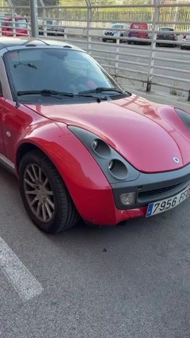 A red Smart car is parked on an asphalt surface with white lane markings. The car has a black convertible top and silver trim. The front of the car is visible on the right side of the frame. A white SUV is parked to the left of the Smart car, also on the asphalt. In the background, there is a metal fence, trees, and buildings. The sky is visible above. The video appears to be a still shot from a higher vantage point, possibly from a drone or elevated position.