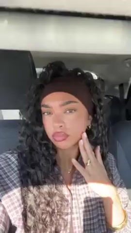 The video shows a young woman with curly dark hair and a brown headband sitting in a car. She is wearing a brown and white plaid shirt and has several pieces of jewelry on her fingers, wrist, and neck. She purses her lips and places her hand near her mouth. The car's interior is visible, including the seats and dashboard. The scene is well-lit.