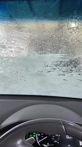 The video shows a car going through a car wash. The perspective is from inside the car, looking out the front windshield. The windshield is covered in soap and water. The steering wheel and dashboard are visible in the foreground. A phone is mounted on the dashboard. The car moves slowly through the car wash.