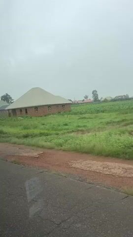 The video shows a cloudy sky over a rural landscape. In the foreground, a dirt road runs alongside a field of tall green grass and some brown soil. In the middle ground, a brick house with a light-colored roof is situated in the field. Further in the background, several other houses with different colored roofs are visible, along with trees and more fields. A person wearing a green rain jacket and carrying a black bag walks away from the camera in the middle ground. The overall scene suggests a residential area in a developing or rural setting, possibly during or after rain due to the wet appearance of the road and the cloudy sky.