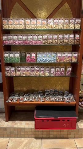 The video shows a display of confections in a shop. There are four wooden shelves with glass fronts, filled with assorted candies. The shelves are arranged in a U-shape around a central display stand with postcards.  A red plastic crate sits on the floor below the candy shelves. The floor is tiled in an earth-tone pattern, and a chandelier hangs from the ceiling. Additional confections are shown in shelves to the side.