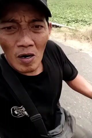 A close-up, first-person perspective video shows a man wearing a black t-shirt and a black baseball cap. He is on a motorcycle, holding the handlebars with his right hand. The man is looking at the camera and speaking. He has facial hair and appears to be outdoors in a rural or suburban area, with a road and vegetation visible in the background. The video is shaky, suggesting movement.