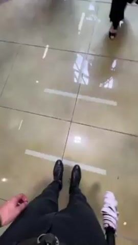 The video shows a first-person perspective inside what appears to be a train station. The camera focuses on the floor, a light yellow tile with dark grout lines and white lines marking areas to stand. Several people walk around, some visible only from the waist down. The camera then pans upward, showing automatic ticket gates and people walking through them. Overhead signs in both Japanese and English are visible, indicating directions. The environment is bright and well-lit with overhead fluorescent lighting.