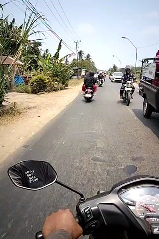 The video shows a first-person perspective of someone riding a motorcycle on a paved road. The rider's hand is visible on the handlebars, and the motorcycle's speedometer is in the foreground. Several other motorcycles, some with passengers, are traveling in the same direction ahead of the rider. A white van is also visible further up the road. The environment appears to be a rural or semi-urban area with houses and vegetation lining the road. The sky is clear and the sun is shining, indicating it is daytime. The rider is wearing a helmet, and the other motorcyclists are also wearing helmets and protective gear. The overall scene depicts a group of people riding motorcycles together.