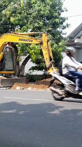 An excavator is digging a hole on the side of a road. A motorcycle speeds past the scene.