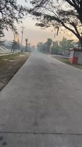 This video shows a first-person view of a person riding a motorcycle down a paved road in what appears to be a rural or semi-rural area during the early morning. The sky is hazy and shows signs of sunrise or sunset. On either side of the road, there are power lines supported by wooden poles, and some scattered trees and vegetation. On the left side of the road, there is a large advertising billboard structure. On the right side of the road, there are signs of construction or landscaping, with piles of gravel and some machinery visible, as well as some basic structures that could be houses or sheds. The road itself is mostly clear of other vehicles, and the overall atmosphere is calm and quiet. The video captures the motion of the motorcycle, with occasional blurring due to movement. The rider's hands or part of the motorcycle's handlebars are visible in the foreground at times, indicating the perspective of the rider.