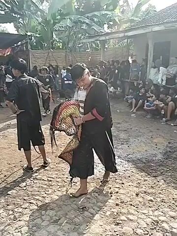The video shows a group of people gathered in an outdoor space, likely a village or rural setting. Several individuals are wearing traditional clothing, and some are carrying what appear to be ceremonial masks or props. The environment consists of a dirt ground, with trees and foliage surrounding the area. There is a visible structure or tent-like covering in the background, and a crowd of onlookers is present. The overall scene suggests a cultural event or performance taking place in a community setting. Some individuals are walking around, possibly preparing for or participating in the event.