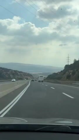 The video shows a point-of-view shot from inside a car traveling on a highway. The highway has multiple lanes and is bordered by hills on either side. The sky is cloudy with patches of blue visible. Several cars and a bus are visible on the highway ahead. Power lines and a utility pole are also visible in the distance. The car's dashboard is partially visible in the foreground. The overall impression is of a daytime journey on a scenic route.