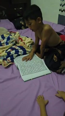 A young boy, shirtless and wearing patterned pants, sits on a bed with a purple sheet. He is reading an open book, likely religious text, with his finger pointing to a line of text. Next to him is a pile of blankets. Another child's hands are visible at the bottom of the frame, also on the bed. The background shows part of a wall with a curtain and a wooden headboard. The lighting appears to be indoor.