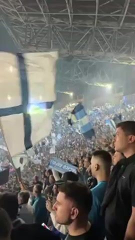 Here is a concise description of the video:

The video shows a large crowd of soccer fans, mostly dressed in blue and white, waving flags in a stadium. The atmosphere is energetic, with smoke or mist filling the air, illuminated by stadium lights. The view is from within the crowd, focusing on the fans, their flags, and a glimpse of the soccer field.