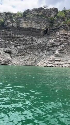 The video shows two small red boats traveling on a body of water with green-tinted water.  The background features steep, layered rock formations along the shoreline. The sky is overcast with scattered clouds. The boats appear to be moving away from the camera's point of view, further into the water and between the rock formations.