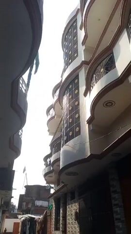 The video shows a narrow street between buildings in what appears to be a residential area. The building on the right has multiple stories, with rounded balconies and decorative metalwork on the windows. The building on the left is simpler in design, with a brick and plaster facade. Two motorcycles are parked on the street. A child is partially visible looking out from behind a brick wall on the left. Further down the street, an adult in red clothing is walking. A Pakistani flag is visible atop a building at the end of the street. The sky is bright, suggesting it is daytime.