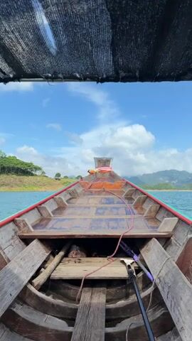 The video shows a first-person perspective from a wooden boat traveling on a body of water. The boat has a canopy providing shade, which is visible in the foreground. The boat itself is made of weathered wood with a red and green trim along the sides. There are wooden planks and some ropes inside the boat. The water is a calm blue-green color. In the distance, there are lush green hills and mountains. The sky is blue with scattered white clouds. The boat appears to be moving forward, indicated by the perspective shifting slightly. There are no people clearly visible in the frame, though a hint of someone in the front of the boat is visible at 00:01.