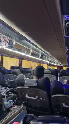 The video shows the interior of a bus with passengers seated. The lighting is dim, with blue and white LED lights illuminating the aisle and overhead compartments. Luggage is stored in the overhead bins. The bus appears to be moving, as indicated by the blur of the exterior through the windows. The camera pans from the interior to the exterior, revealing a man in an orange shirt and blue shorts standing outside the bus. The exterior environment is dimly lit with streetlights and greenery.