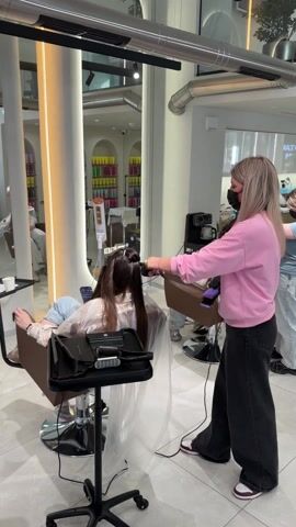The video depicts a salon scene with two hairstylists attending to clients. On the left, a client with long brown hair is seated in a salon chair, draped with a clear plastic cape. A stylist wearing a pink sweater and black pants, also masked, is straightening the client's hair using a flat iron. The stylist holds the client's hair taut with one hand while running the flat iron down the strand with the other. A small cart with a black surface stands beside the client, holding various items.

In the background, another stylist, wearing a blue shirt and light-colored jeans, is tending to a second client near a sink area. The environment is well-lit and clean, featuring a modern interior design with white walls, tiled flooring, and sleek metal fixtures. A large mirror on the left reflects a shelving unit filled with colorful hair product bottles. Other salon equipment, such as a coffee maker, can be seen in the background, contributing to the professional setting.