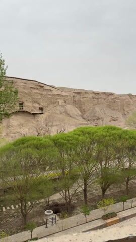 From a vantage point with a light brown wooden railing, the video captures an expansive view of a desert landscape featuring cave dwellings built into a hillside. The hillside is a sandy beige color. Below, a paved walkway lined with trees, primarily green in color, leads through the area. Several small, tan-colored buildings with distinct roof shapes stand along the walkway. People can be seen strolling along the walkway. The sky is overcast, creating a muted, grey light.