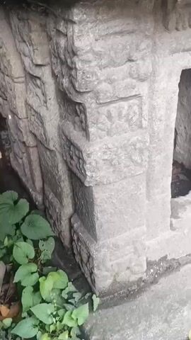 This video shows a close-up of an old stone structure, possibly a shrine or monument, covered in moss. The stone is intricately carved with various patterns and designs. The structure appears to be outdoors, with some green foliage visible in the lower left corner. The lighting suggests it is daytime.