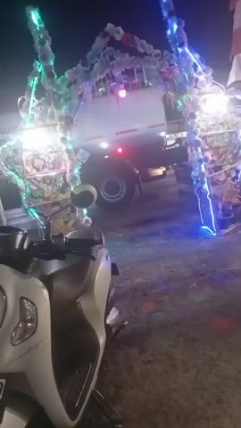 The video captures a nighttime scene where a white scooter is parked in the foreground. Behind the scooter, an archway decorated with colorful lights and flowers spans across an area. A white truck with its brake lights illuminated is visible passing through or parked beyond the archway. The environment appears to be an outdoor space, possibly a street or a venue, with the ground being dirt or gravel. The lighting is predominantly from the decorative lights on the archway and the truck.