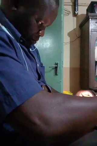 A medium indoor shot shows a dark-skinned man wearing a blue button-down shirt and white earbuds, concentrating intently on a task at hand. His hands are brightly illuminated, suggesting a light source is directed at his work. In the background, a pale green door with a silver handle and a dark wooden cabinet with glass paneling are visible. A clothes hanger is on the wall in the upper-right. The man's posture and focus suggest he is engaged in a detailed, manual activity.