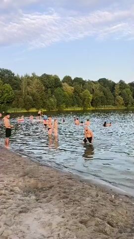 A group of people are enjoying a sunny day at a lake. The camera is positioned on a sandy shore, looking out at the water. In the foreground, the sand is smooth with some small ripples. The water is calm and reflects the sky. Several people are swimming and playing in the water. Some are on inflatable tubes, while others are swimming freely. One person is standing in the water, looking towards the shore. In the background, a dense line of trees forms the edge of the lake. The sky is a light blue with scattered white clouds. The overall atmosphere is one of leisure and recreation.
