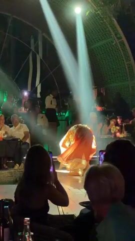 A female belly dancer in a sequined bra, orange skirt and a white shawl performs on a wooden stage in a restaurant, illuminated by white spotlights. The restaurant has a domed ceiling with a green light effect. Patrons at tables around the stage are filming the performance on their cell phones. Some of the staff are looking on. The scene is lively and celebratory.