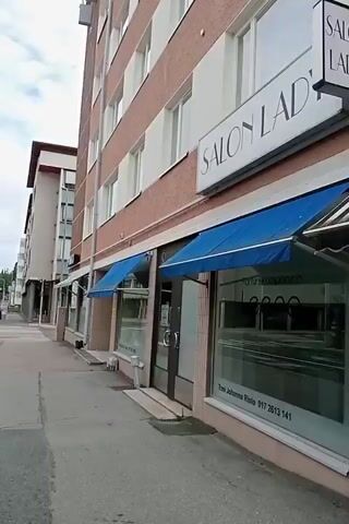This video shows a street view in Kuopio, Finland. The scene is set on a sidewalk next to a street with multiple lanes and a crosswalk. Buildings line the sidewalk, with businesses on the ground floor.

In the first few seconds, a tall building with a red brick facade and white trim is visible, featuring multiple windows. A street lamp and traffic lights are also present. A salon called "SALON R" is advertised on the building's upper floor.

As the video progresses, the camera pans to reveal more of the street and surrounding buildings. A grey, multi-story building with many windows is seen across the street, with a sign that reads "LIMHAT BLOCK" on a pole in the foreground. Further down the street, another building with a yellow facade displays shop windows. Inside one of these windows, mannequins are dressed in formal wear, with text visible reading "JUHLA-ASUKOT" and "FRAKKIKAUppa". Another shop window has text that appears to be "EMOON" or similar. A green awning is visible over one of the shop entrances.

The weather appears to be overcast, suggesting a cloudy day. The time of day is difficult to determine precisely but appears to be daytime, possibly late morning or afternoon, given the lighting. There are no people clearly visible walking or engaging in any specific activity. The overall mood is neutral, presenting a straightforward depiction of an urban street scene.