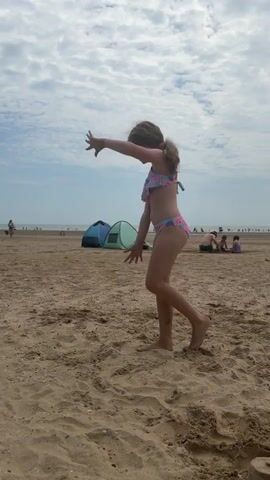 A young girl in a colorful bikini dances on a sandy beach. The beach is populated with other people, tents, and sandcastles, suggesting a bright, sunny day. The sky is partly cloudy, indicating it's likely daytime. The girl is performing a series of graceful movements, including a handstand. Her posture and expression suggest she is enjoying herself and is confident in her abilities. The overall scene is one of carefree fun and activity on a pleasant beach day.