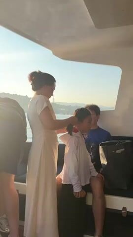 This media file captures a serene moment aboard a boat, likely a ferry or private vessel, in Labuan Bajo, Indonesia. The primary subjects are three individuals: an adult woman, a young girl, and an adult man. The woman, dressed in a long, flowing cream-colored dress, is attentively braiding the young girl's hair. The girl, wearing a light-colored hoodie, sits facing away from the camera, looking out towards the water. The man, in a blue t-shirt, is seated behind the girl, his expression suggesting a relaxed and possibly amused observation of the scene.

The setting is outdoors, on the deck of a boat. The bright sunlight indicates it is daytime, likely late afternoon or early evening, given the warm, golden hue of the light. The sky is clear and blue, suggesting fair weather. In the background, a coastline with buildings and greenery is visible across the water, typical of a harbor or island setting. The overall mood is peaceful and intimate, highlighting a tender moment between family members. The presence of a large black bag on the seating area suggests travel or a day trip. There is no visible text within the frame.