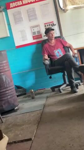 A man wearing a red t-shirt, dark pants, and a cap sits in a black office chair, facing to the right, with his legs crossed. He appears to be in an indoor space with blue walls. Behind him, a bulletin board displays several documents. To his left, a large metal barrel and a black trash bag are visible on the floor. The floor is made of concrete or stone tiles. The man is speaking, though his words are partially obscured by background noise.