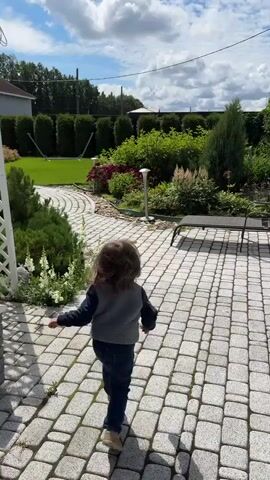 A young child with long brown hair runs away from the camera down a winding cobblestone path in a well-maintained yard. The child wears a grey vest over a dark long-sleeved shirt and blue jeans. The yard features lush green grass, manicured hedges, flowering plants, and a modern house in the background. The sky is bright blue with fluffy white clouds, suggesting a sunny day. The child's movement is energetic, with arms outstretched slightly, as they move further away into the garden.
