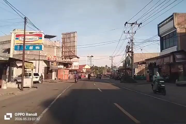 The video shows a view from inside a vehicle traveling down a road in a small town. The road has a clear center line, with buildings and small businesses lining both sides.  The buildings appear to be a mix of one and two stories. There are numerous overhead power lines crisscrossing the sky. Several motorbikes and a few cars are traveling in both directions on the road.  Some small food stalls or shops are visible on the right side of the road. The sky is a light blue. The timestamp on the video indicates the date is May 23, 2026, at 07:32.  The device used to record the video is identified as an OPPO Reno8.