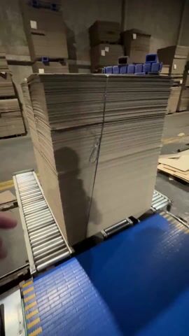 A man's hand with tattoos is briefly seen touching the side of a large stack of cardboard sheets. The cardboard is strapped together and is resting on a roller conveyor belt system in what appears to be a warehouse or industrial setting. The background shows numerous stacks of cardboard and blue crates. The conveyor belt is primarily silver with blue accents. The camera perspective is first-person.