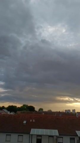 The video shows a view of rooftops and a cloudy sky at sunset. The sky is filled with gray and dark clouds, with patches of yellow and orange light from the setting sun peeking through. The rooftops are mostly made of red tiles, with some houses also having solar panels installed. In the distance, there are some trees and taller buildings visible. The overall atmosphere is calm and serene, with the fading light creating a soft glow over the scene.
