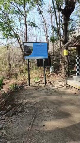 The video begins with a sign on a post in a wooded area. The sign has an image and text. To the right of the sign is a small, open-sided structure with a tiled roof and checkered pillars. Two people are sitting on a concrete platform beneath this structure. One person is wearing a hijab and a red and white top, and the other is wearing a dark jacket and pants. A motorcycle helmet is visible on the platform. The ground is dry and dusty, with sparse vegetation. The camera pans to the right, revealing more of the wooded environment and a rocky landscape. The sun is casting long shadows.