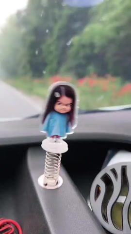 A close-up shot inside a vehicle shows a small, acrylic bobblehead-style figure of a cartoon girl with long dark hair, a light blue dress, and an angry expression, mounted on a spring on the dashboard. The figure bounces slightly as the vehicle moves. Through the windshield, a road with a green, tree-lined shoulder is visible. To the right of the figure, part of a circular car air freshener with a silver, patterned grille is seen. To the left, a coiled red and black cable is partially visible.