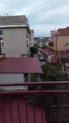The video shows an outdoor view of a neighborhood street, filmed from a balcony. The sky is overcast. Various buildings are visible, primarily houses and apartments with red or gray roofs. Several cars are parked along the street. Trees and greenery are interspersed among the buildings. The balcony has a red metal railing. The camera pans slightly to the right during the video. A voice can be heard speaking in the background, possibly in Russian.