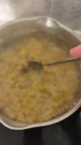 A pot of macaroni is being stirred with a spoon in boiling water. The camera is positioned directly above the pot, and the steam from the water obscures the view of the macaroni. A hand is visible holding the spoon and stirring the macaroni. The pot is on a black stovetop, and the background is blurred.