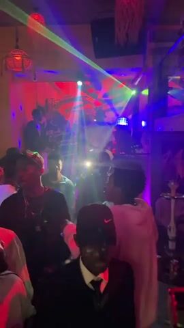 The video captures an indoor party or club scene filled with numerous young adults dancing and socializing. A DJ, wearing a light-colored shirt and a cap, stands behind a console in the background, actively playing music. The room is dimly lit, illuminated primarily by dynamic laser lights casting red, green, and blue beams across the space. The walls appear to be a light brown or yellow color, and several decorative, woven lamps hang from the ceiling. In the foreground, a person wearing a dark suit, tie, dark cap, and dark sunglasses is visible. Another individual in the mid-ground wears a white shirt and white-framed sunglasses, while a person on the right wears a light-colored hoodie. Many attendees are holding up smartphones, some with their flashlights on, contributing to the ambient light. Hookahs are visible on a table on the far right side of the frame. The overall atmosphere is energetic and lively.