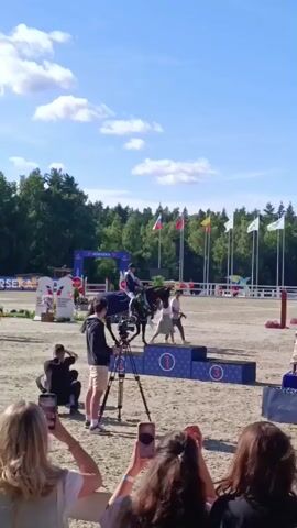 A horse and rider are on a podium. The horse is decorated with a wreath of flowers and a blue blanket. The rider is wearing a dark helmet, a light blue jacket, and white pants. Two people, a man and a woman, are walking away from the podium. A man in a suit is standing next to a table with trophies and awards. In the background, there are flags and trees. A camera crew is filming the event. A crowd of people are watching from the foreground. The sky is blue and there are some clouds. The ground is made of sand.