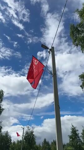 The video shows a red flag with a white hammer and sickle blowing in the wind, attached to a light post. The flag is partially obscured, and the text on it is not clearly visible. The sky is blue with scattered white clouds. The light post is grey and has a street lamp attached to it. There are trees visible on either side of the frame, and a sidewalk runs alongside a grassy area. In the background, another flag is visible on a distant light post. Further back, a blue and white structure resembling a playground is present. The camera pans slightly up and down.