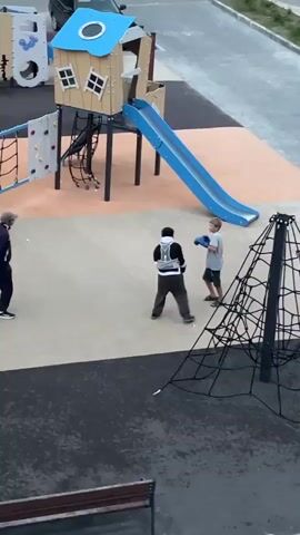 Four boys are on a playground. Two boys are wearing boxing gloves, one is wearing a white shirt and dark shorts, and the other is wearing a dark shirt with a vest over it and dark pants. They appear to be sparring. A third boy in a black shirt and pants stands to the left, watching. A fourth boy in a dark tracksuit walks across the background, away from the sparring boys. A blue slide and a climbing structure are visible behind the boys.