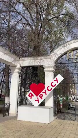 The video shows the entrance to a park. The entrance consists of a white, arched structure with the words "ПАРК семейный" in gold lettering above a black metal gate with decorative swirls. To the left of the gate is a white sign with a red heart and the text "Я УРУССУ". The park inside features benches and trees. The sky is overcast.  The video is taken from a stationary position and pans slightly upwards to show the sign and entrance in more detail.