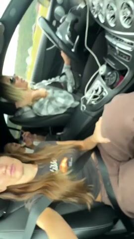 Two young women are in the front seats of a car. The woman in the passenger seat, wearing sunglasses and a grey and white plaid shirt over a black tank top, is speaking with her mouth open and her left hand touching her head. The woman in the driver's seat, with long brown hair and wearing a grey t-shirt with a croissant graphic and a brown sports bra, is looking at the camera and talking. She is wearing a seatbelt. The car's interior is visible, including the steering wheel, dashboard, and center console. The environment outside the car appears to be rural, with green hills in the background. The video is shot from a low angle, looking up at the women.
