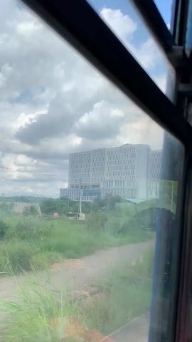 The view from inside a moving vehicle looks out onto a scene featuring a bus stop and a large, modern building in the background. The bus stop has a covered area with benches, and several people appear to be sitting or waiting there. A small blue booth or structure stands next to the bus stop, and several advertisements or signs are displayed nearby. The road in the foreground is unpaved and lined with green vegetation. The large building in the background appears to be multi-story, with a repetitive pattern of windows. The sky is partly cloudy, with patches of blue visible. The frame of the vehicle's window obscures parts of the view, adding a sense of being in transit.